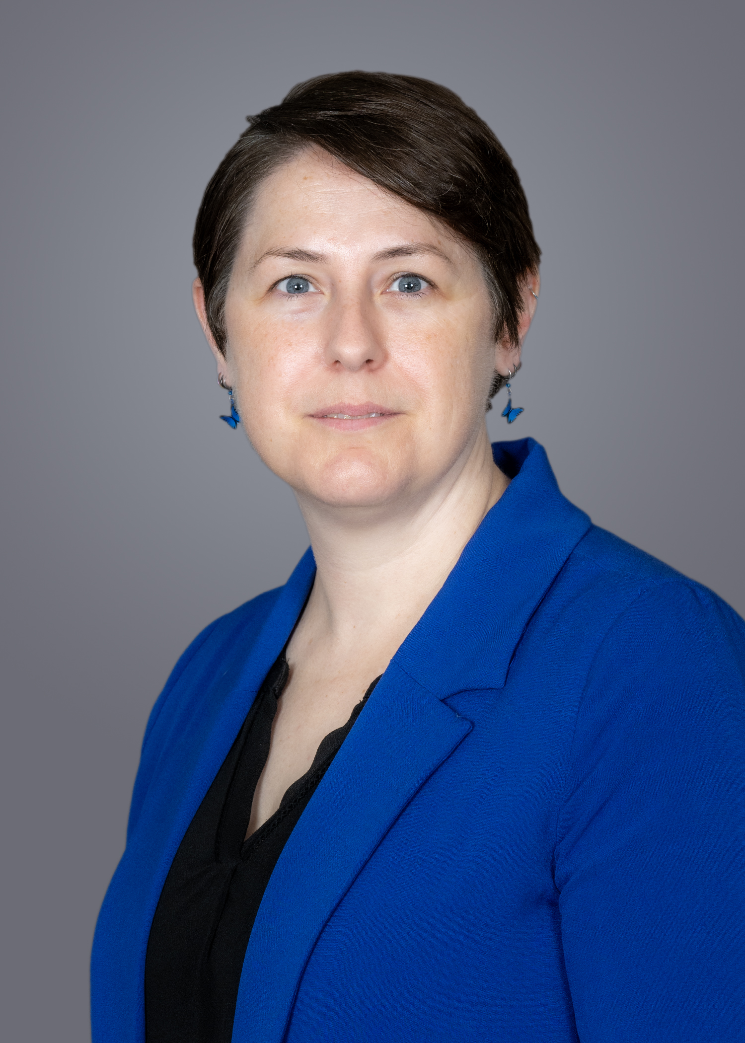 Portrait of a woman wearing a blue blazer and black top, facing the camera with a neutral expression against a gray background.