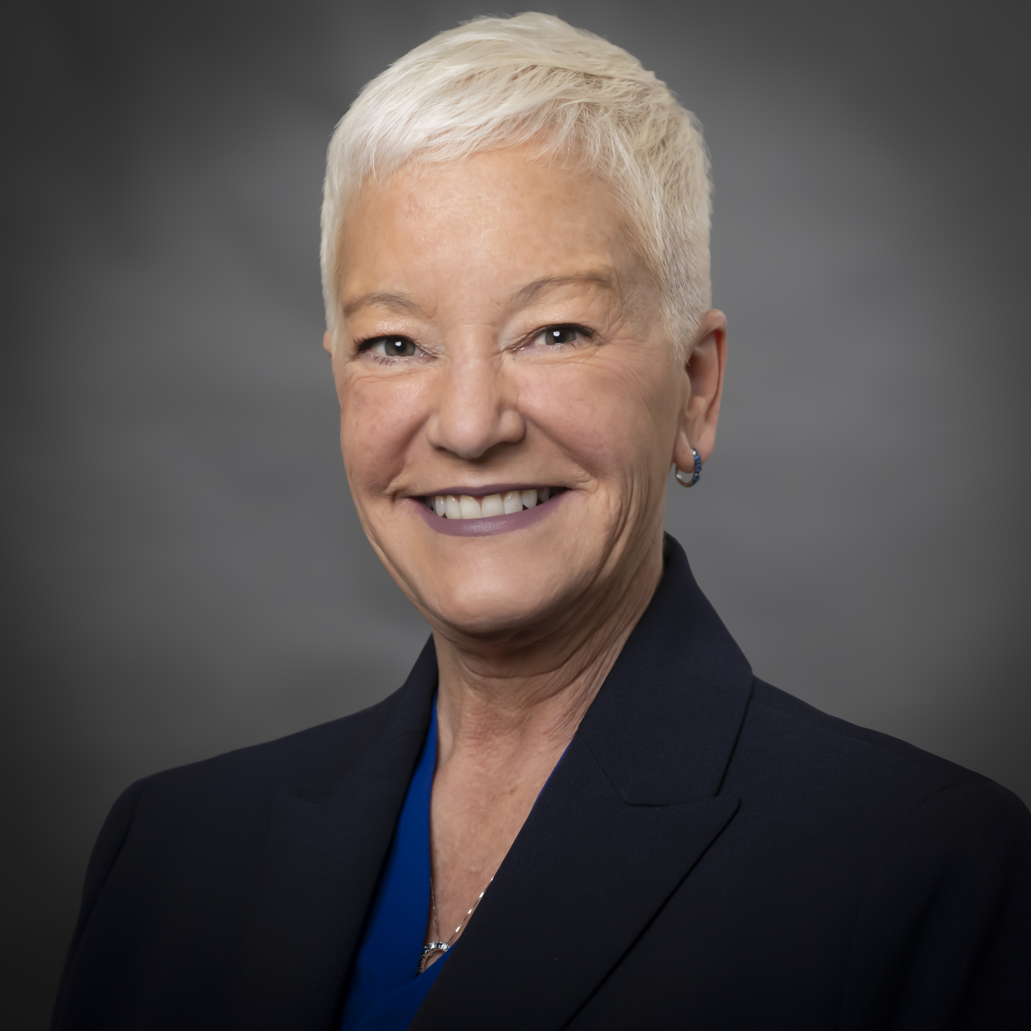 Portrait of a woman with short white hair smiling, wearing a dark blazer and blue top against a gray background.