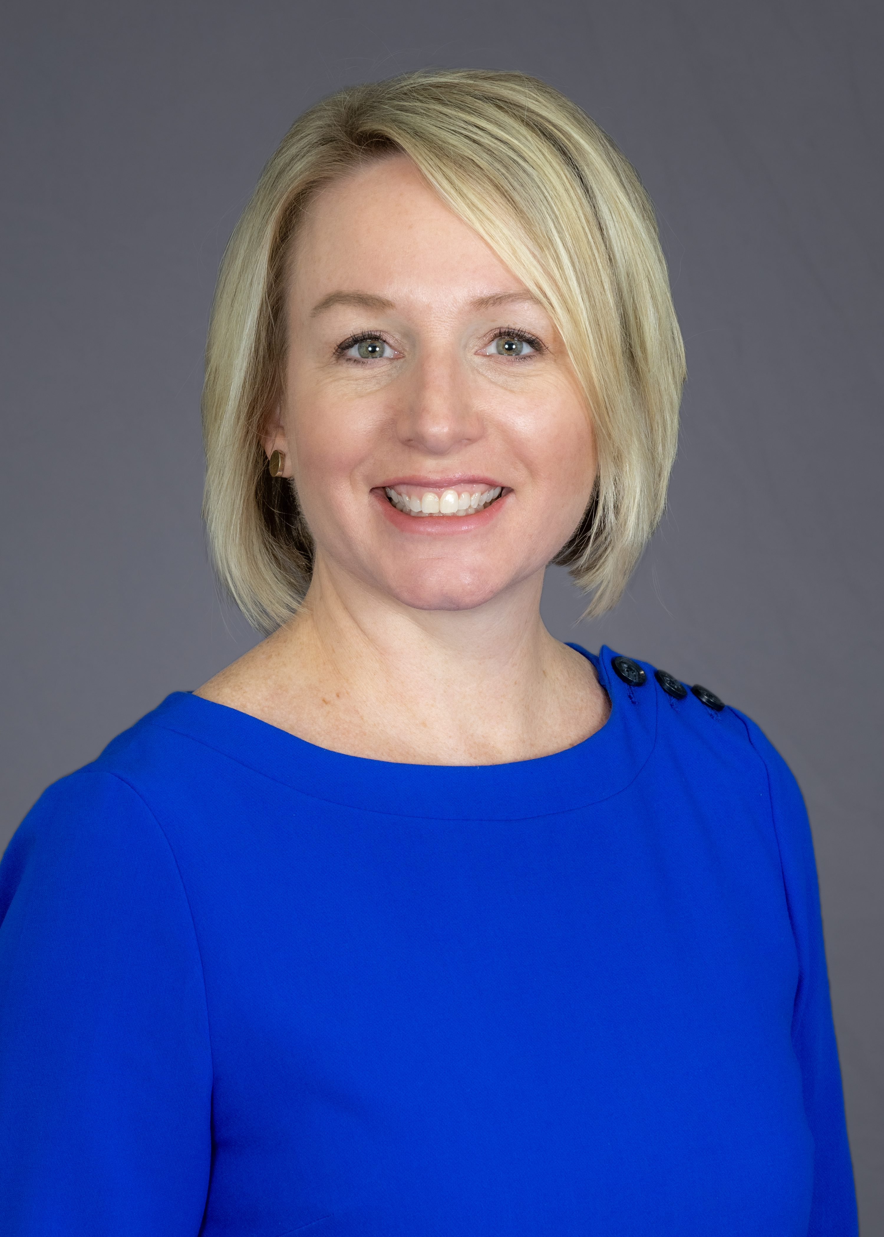 Portrait of a woman with blonde hair smiling, wearing a blue dress against a gray background.