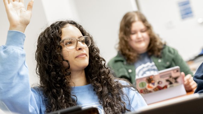 A student wearing glasses raises her hand in a classroom while another student sits behind her using a tablet.