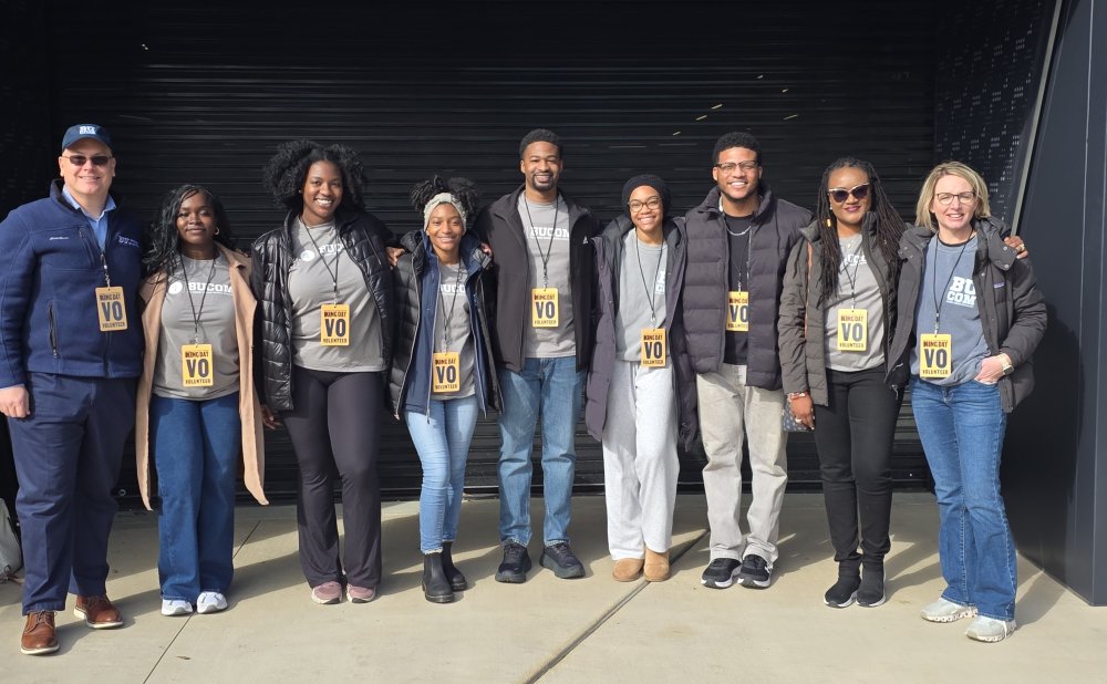 Group of ten adults standing side by side outdoors, smiling at the camera. Most wear gray BUCOM T-shirts layered with jackets and ‘King Day Volunteer’ badges on lanyards. They pose with arms around each other in front of a dark building entrance, suggesting a team volunteer event.