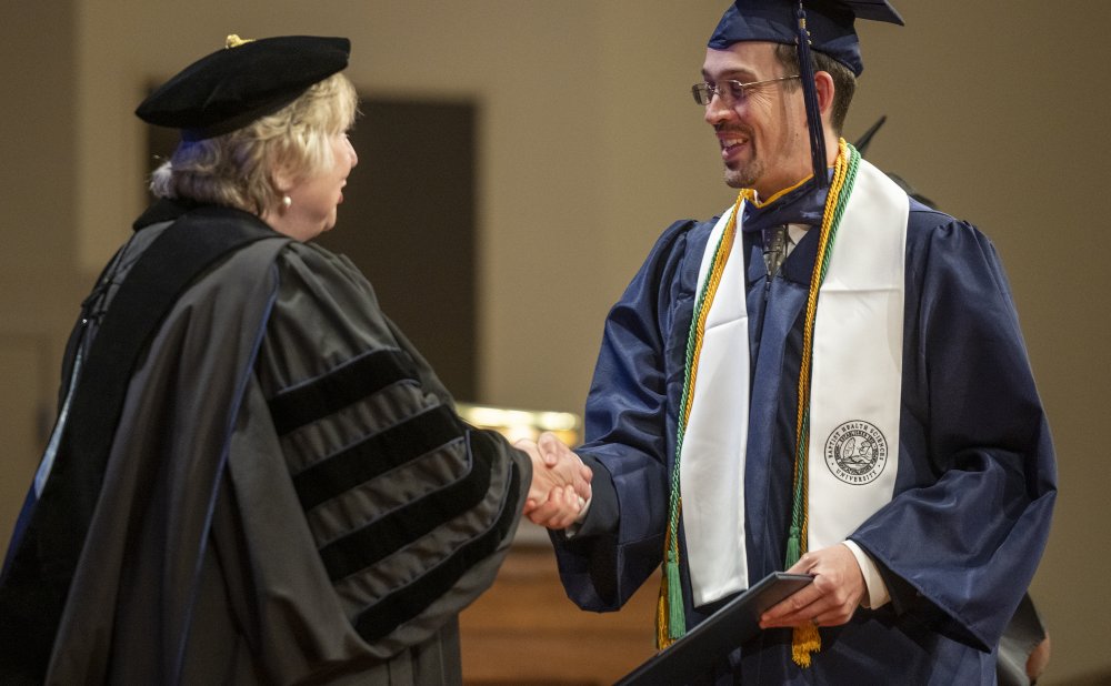 A graduate in a navy blue cap and gown shakes hands with a faculty member in academic regalia during a graduation ceremony. The graduate is smiling and wearing a white stole with a university seal, along with green and gold honor cords. The faculty member is dressed in a black gown with velvet stripes and a traditional academic cap.