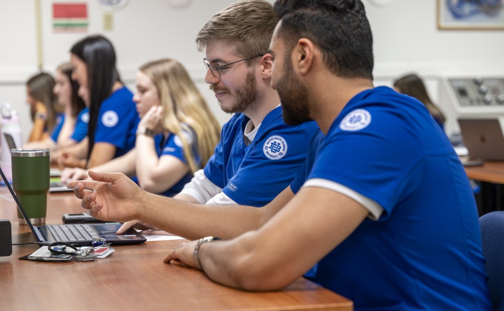 two male students in blue scrubs with a Baptist University patch on their arms looking at a computer