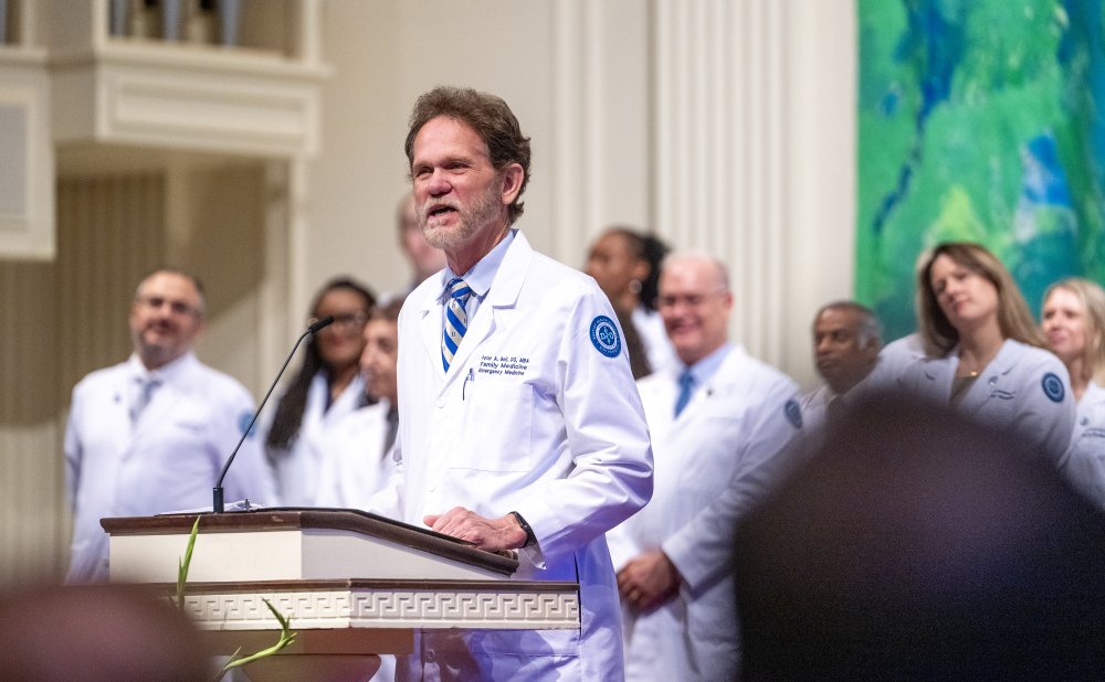 A man in a white lab coat speaks at a podium with a microphone, addressing an audience. Several other individuals in white coats stand behind him on a stage inside a large, well-lit hall.