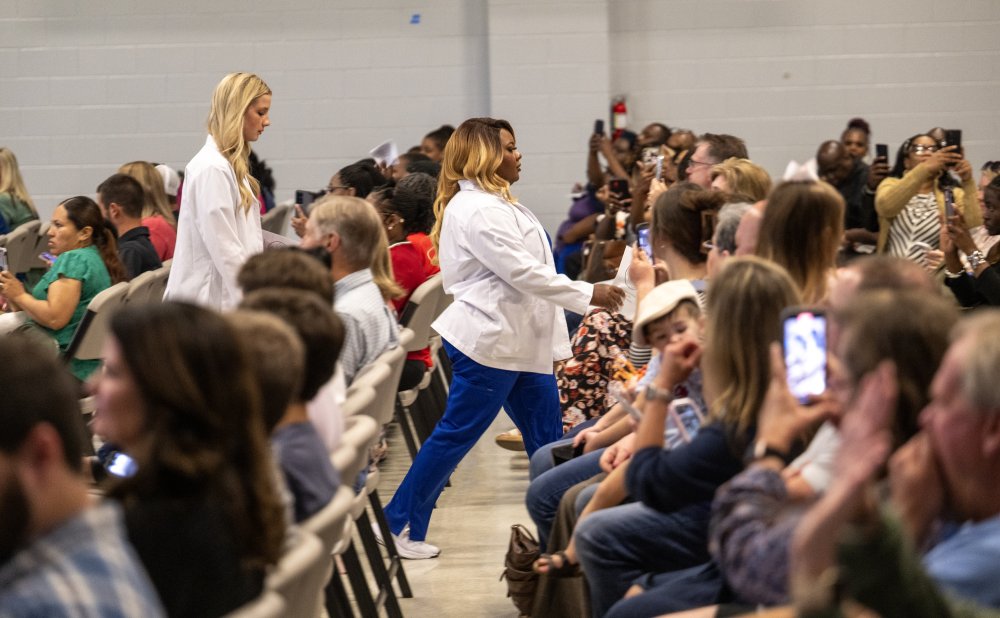Two students in white coats walk down an aisle during a ceremony as family and friends seated on both sides take photos and cheer in support.