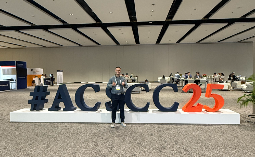 Jonathan Hawkins stands smiling and giving a thumbs-up in front of a large “#ACSCC25” display at the American College of Surgeons Clinical Congress. The conference exhibit hall is spacious with high ceilings, carpeted floors and attendees in the background.