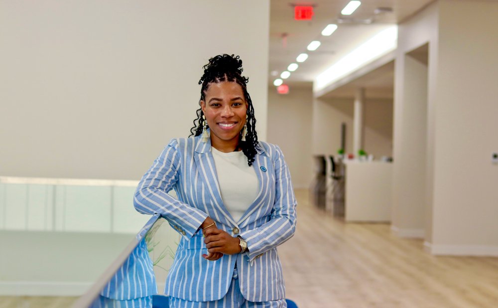 A smiling woman stands confidently in a modern hallway. She is wearing a light blue and white striped suit with a white shirt. Her hair is styled in curls and she is leaning casually against a glass railing. The background features light-colored walls, wooden floors, and a bright, open office space.