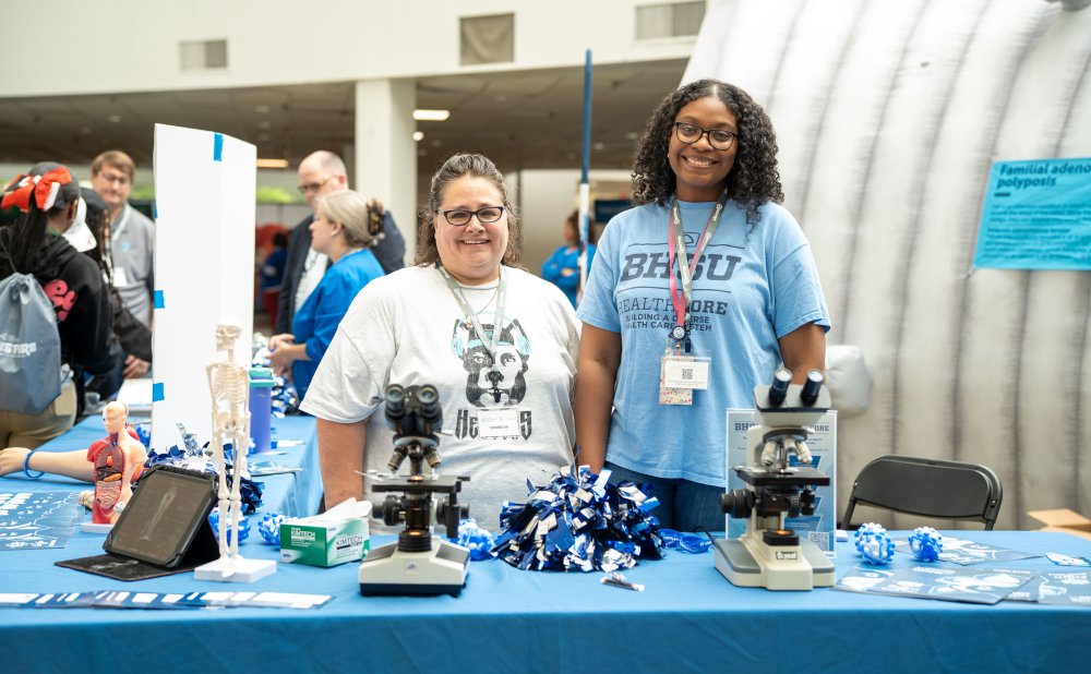 Two women stand behind a BHSU Healthcare booth, smiling at the camera. The table features anatomical models, microscopes, pom-poms, and brochures. One woman wears a BHSU Healthcare t-shirt, and the other wears a shirt with a stylized dog graphic. Behind them, a large inflatable colon exhibit is partially visible.