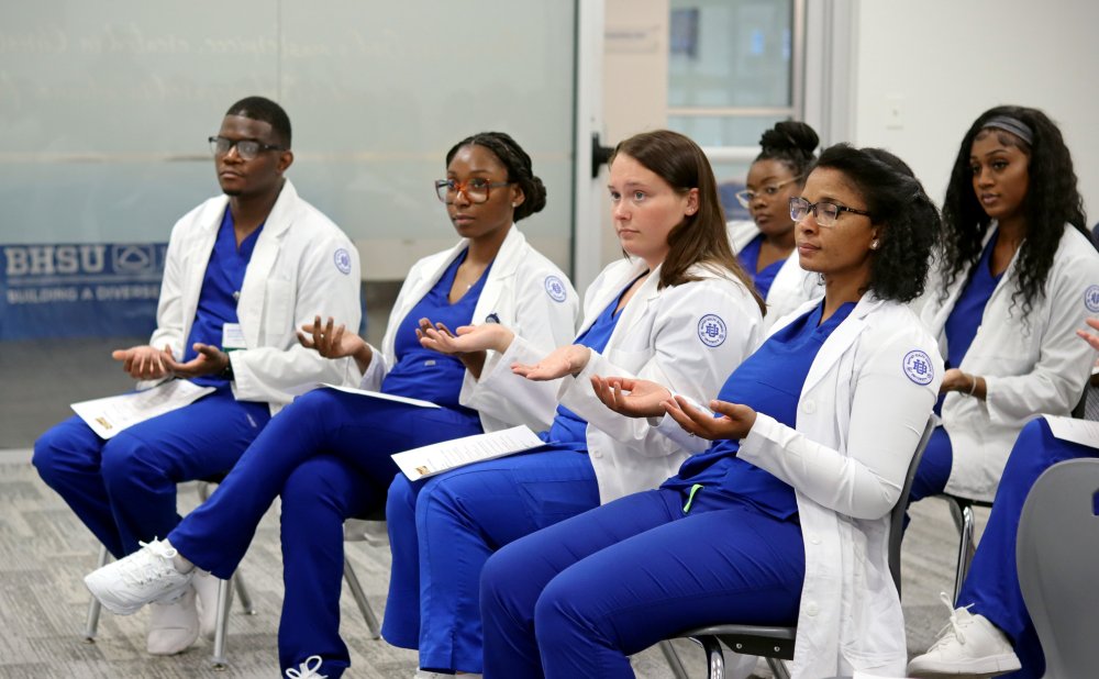 Students dressed in blue scrubs and white coats hold their hands up and out