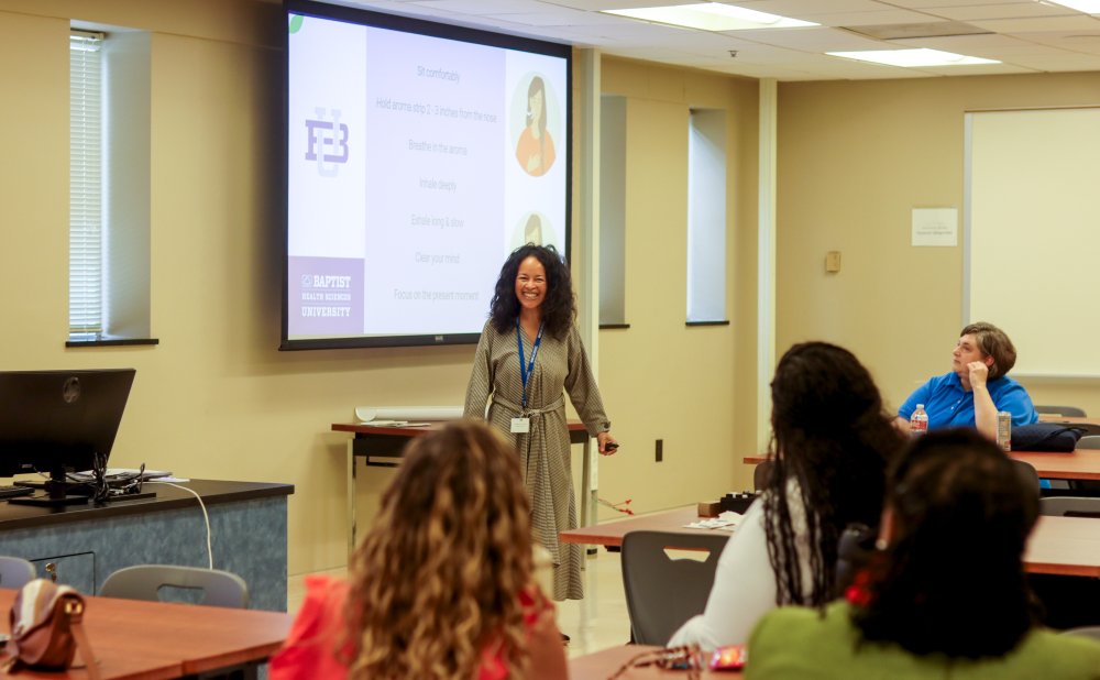 A smiling woman stands at the front of a classroom leading a session, with a presentation slide behind her displaying breathing and relaxation instructions. Participants seated at tables look on with interest.