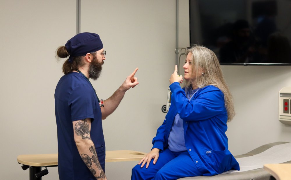 A DNP student in navy scrubs performs a neurological exam on another person wearing royal blue scrubs in a clinical skills lab. Both individuals are holding up one finger while maintaining eye contact to test coordination and focus.