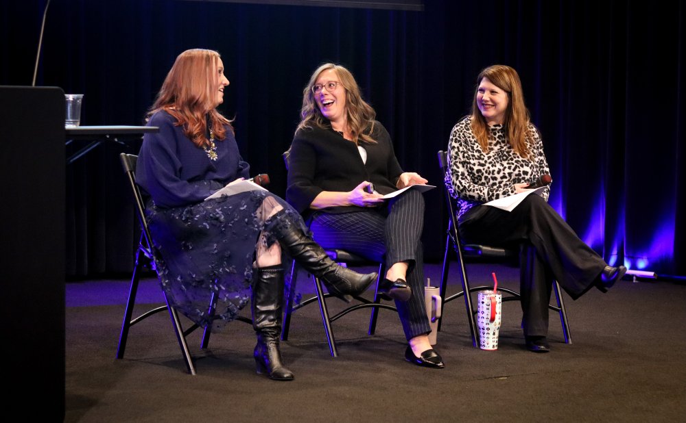 Three women sit on stage in folding chairs during a campus professional development session. They are smiling and engaged in conversation while holding papers. A dark curtain and blue stage lighting are visible in the background.
