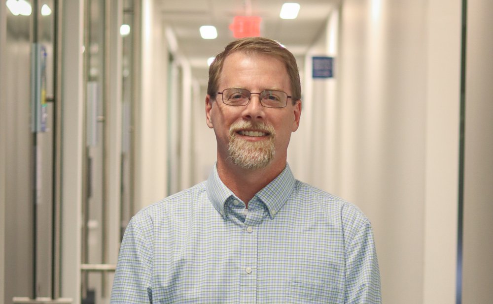 A university faculty member wearing glasses and a light blue checkered shirt smiles while standing in a brightly lit hallway of campus offices.