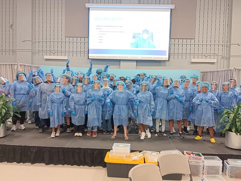 A large group of students stand on a stage wearing full personal protective equipment (PPE), including blue gowns, face shields, gloves, and hair covers. They pose for a group photo, some raising their gloved hands in the air. A presentation slide about PPE is projected on the screen behind them in a classroom or conference setting.