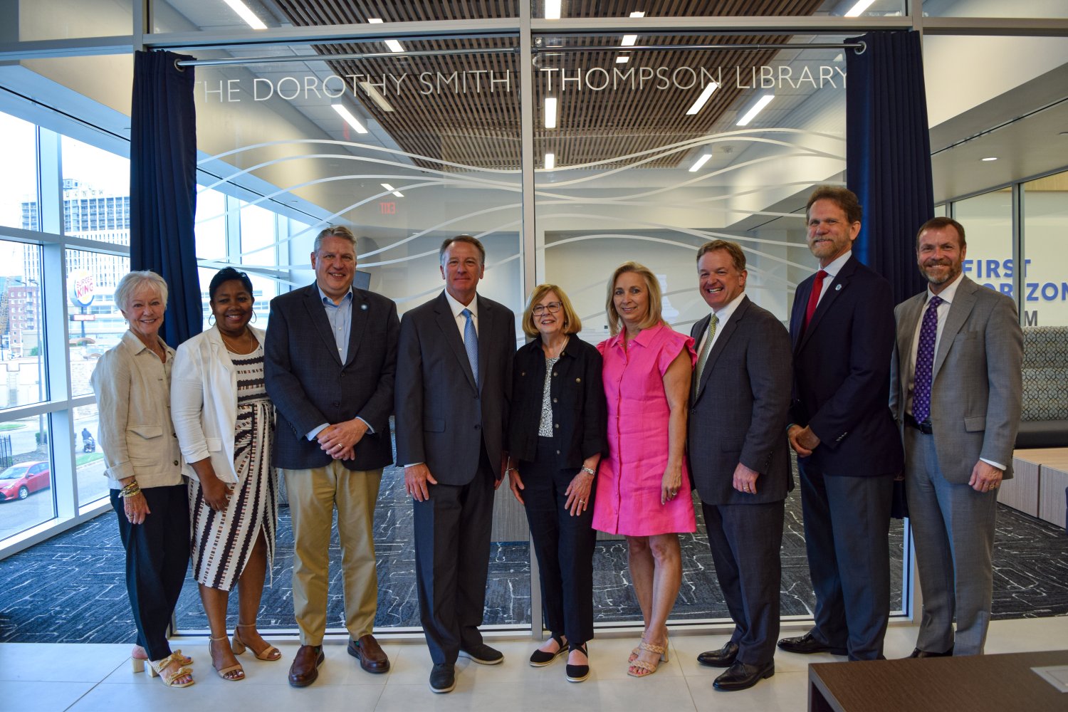 A group of nine individuals stands in front of a glass wall labeled "The Dorothy Smith Thompson Library." They are smiling and dressed in professional attire, gathered for what appears to be a library dedication ceremony. Blue curtains are pulled back on either side of the glass.