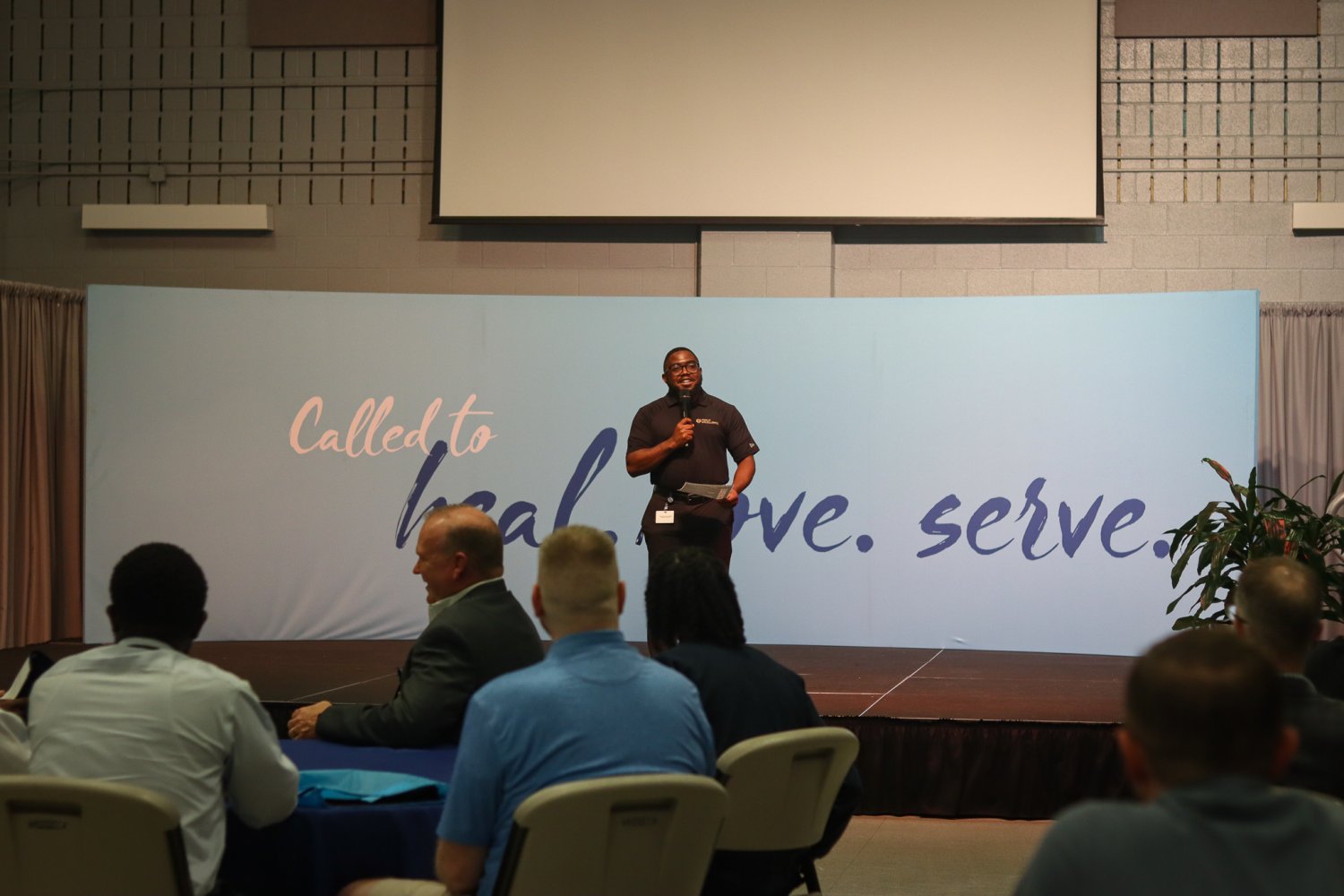 A speaker addressing the audience from stage in front of a backdrop reading “Called to heal. love. serve.” at the networking event.