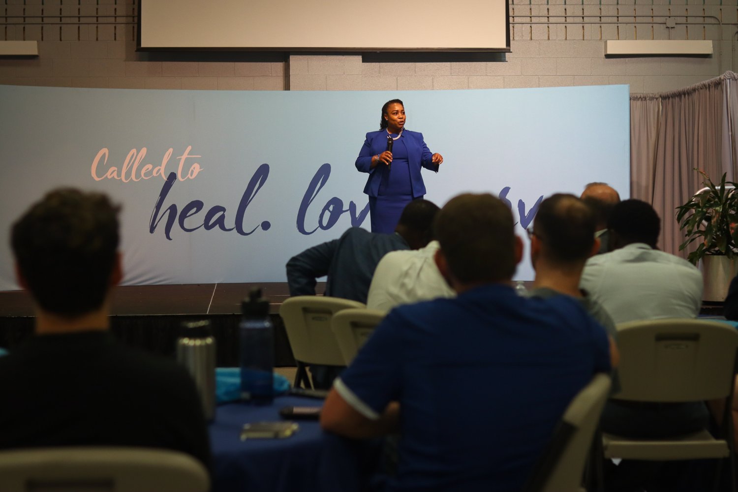 A faculty member in a blue suit speaking to the audience on stage at the Future Men in Healthcare Networking Event.