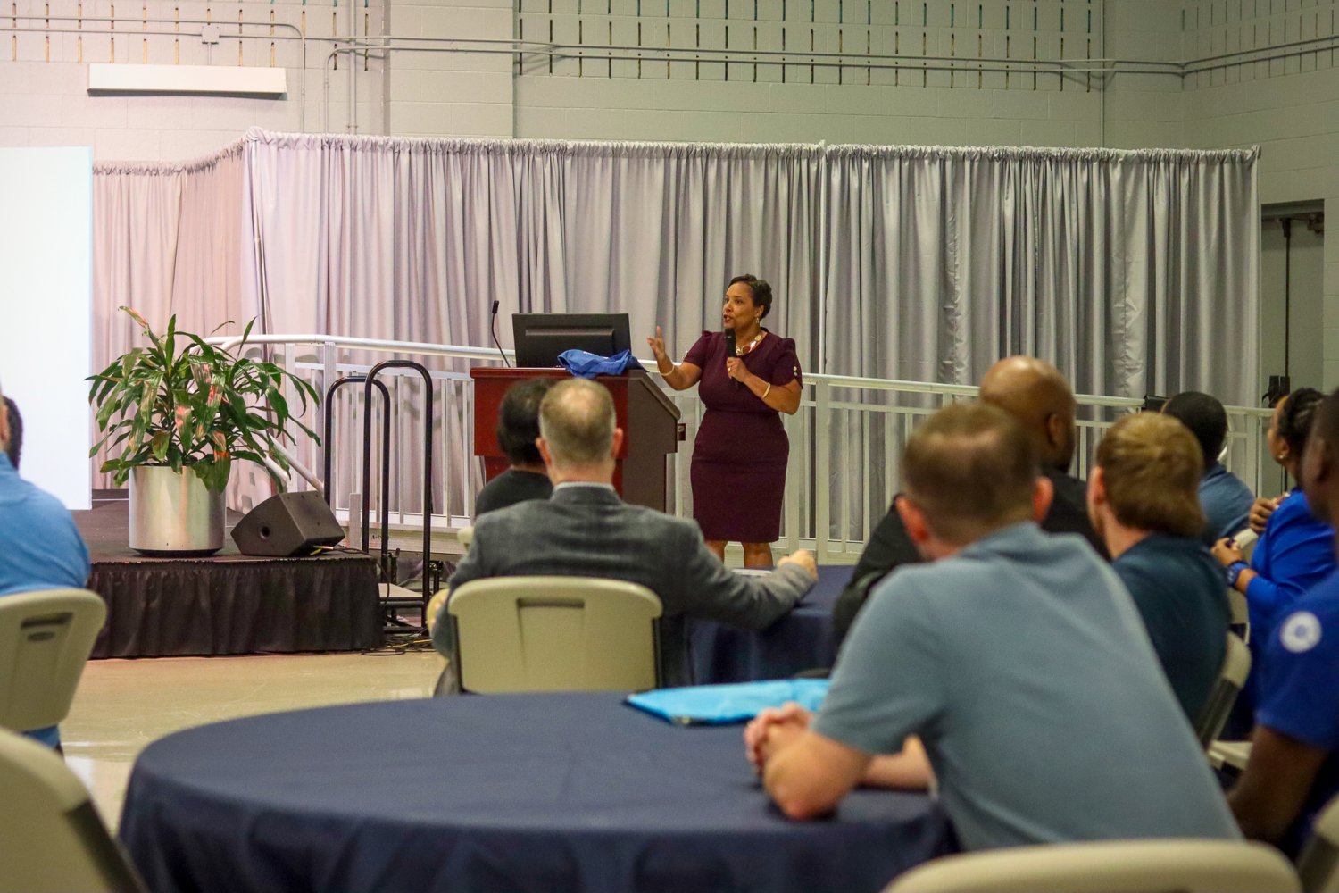 A woman presenting on stage, engaging with attendees seated at round tables during the event.
