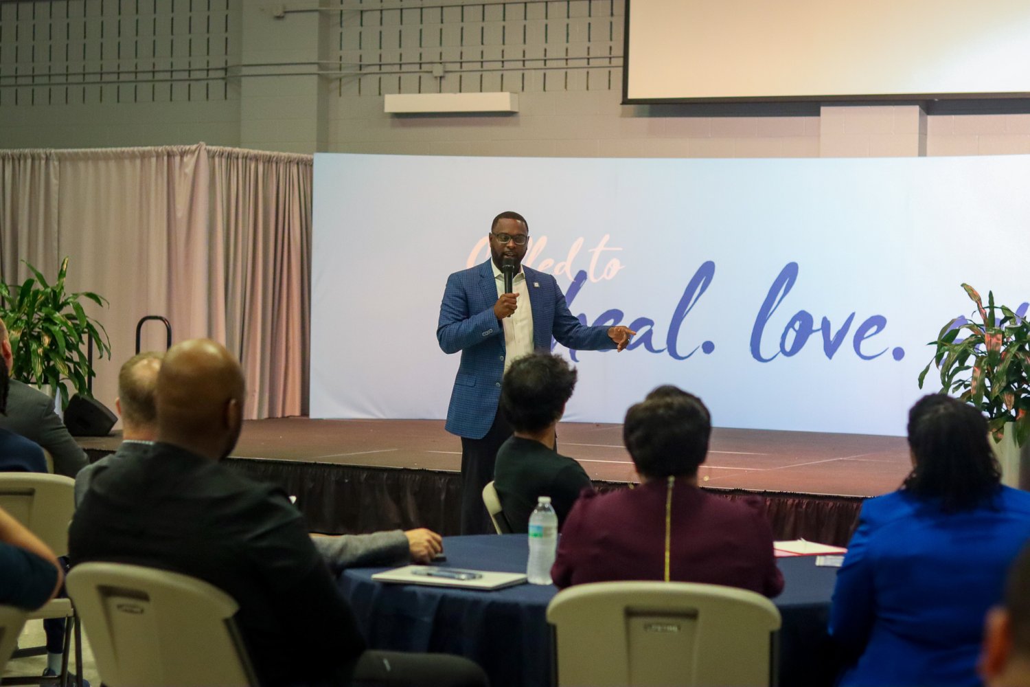 Mayor Paul Young speaking on stage in front of a banner that reads “Called to heal. love. serve.” at Baptist Health Sciences University.