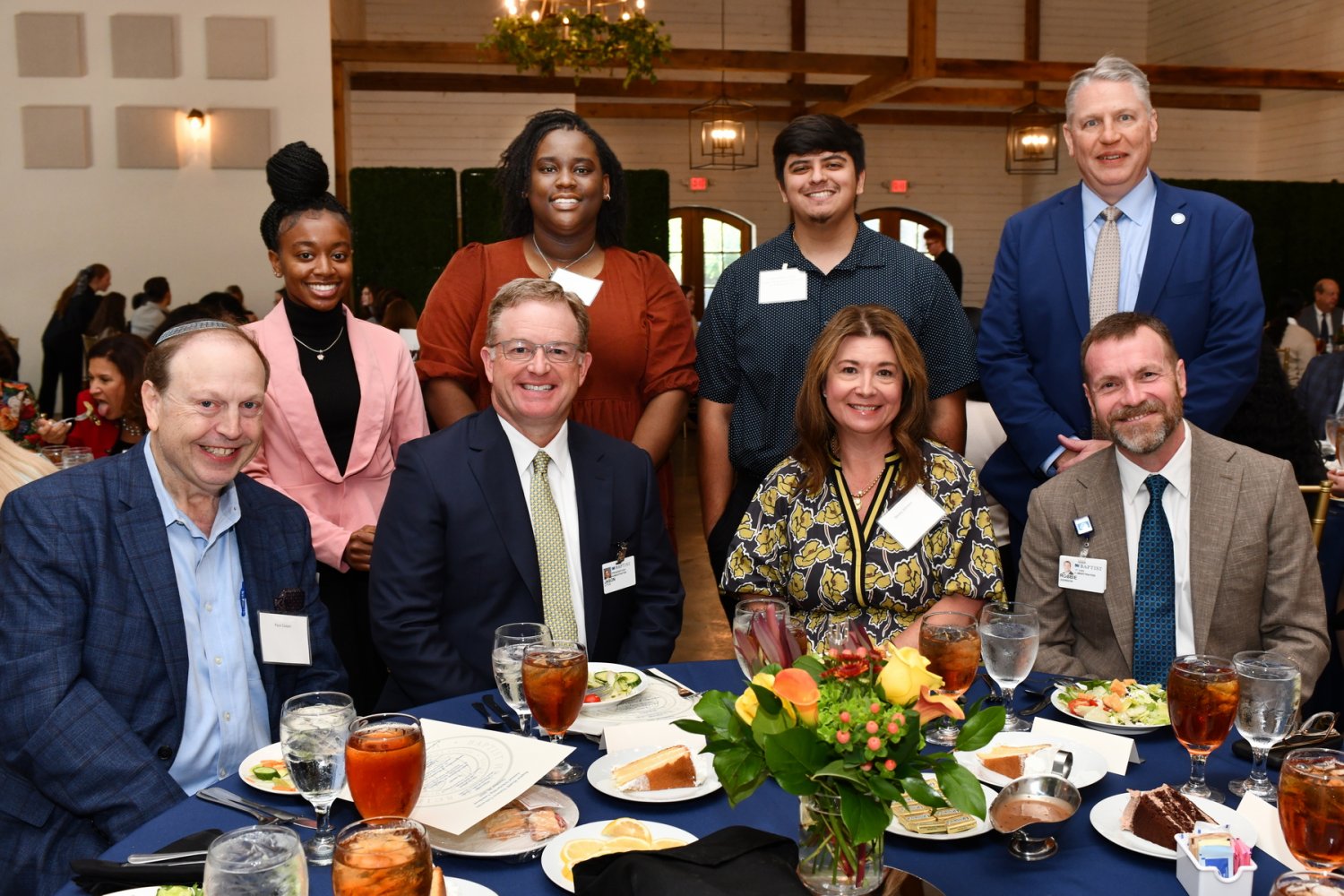 A group of eight people, including students and university leaders, smile around a dinner table at a formal event.