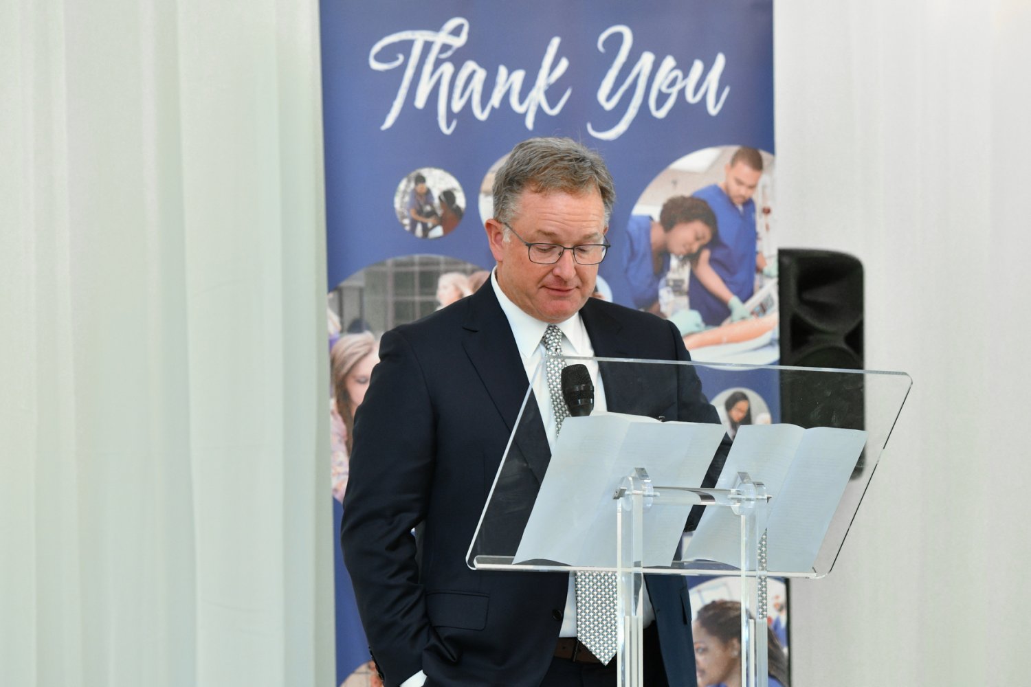 CEO of Baptist, Jason Little, in a suit speaks at a podium with a microphone in front of a “Thank You” banner.