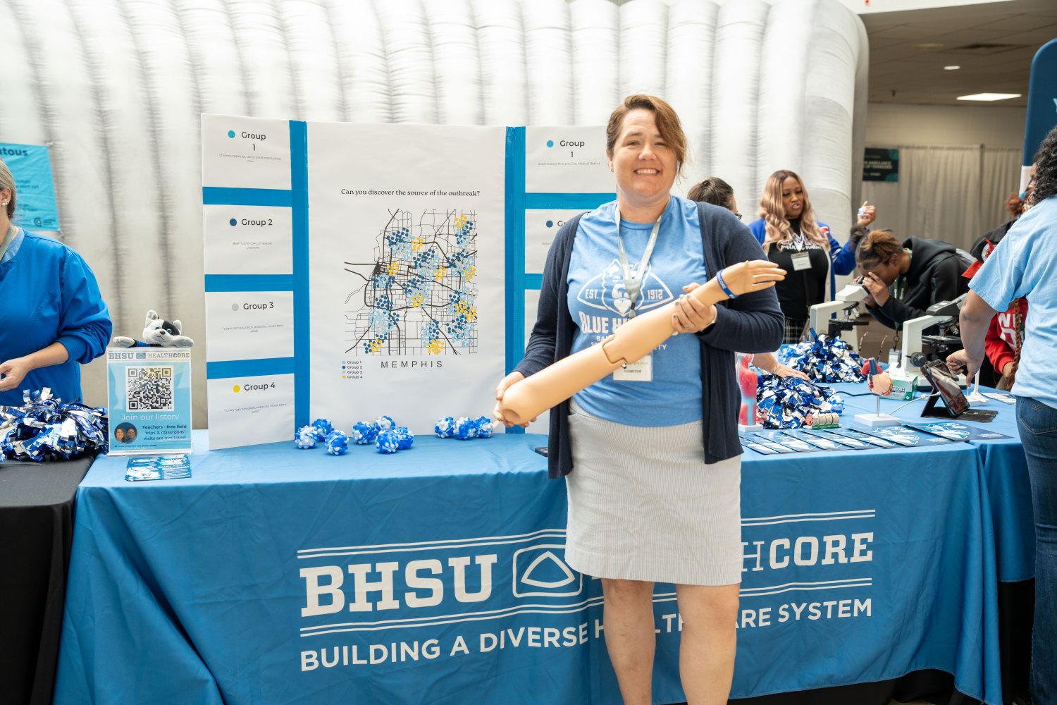 A woman stands smiling in front of a healthcare education booth, holding a medical training prosthetic arm. Behind her is a display board featuring a map of Memphis with colored data points and group labels, asking “Can you discover the source of the outbreak?” The booth is covered with a blue tablecloth labeled “BHSU Healthcare – Building a Diverse Healthcare System.” Other people are interacting with microscopes and informational materials at the booth. The setting appears to be a community health or educ
