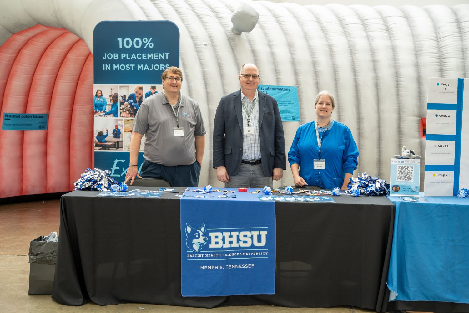 Three individuals stand behind a BHSU (Baptist Health Sciences University) booth at an event. The table is covered in promotional materials, pamphlets, and blue-and-white pom-poms. Behind them is a large inflatable structure resembling the inside of a colon, along with a banner promoting "100% job placement in most majors." A sign on the table reads “BHSU – Memphis, Tennessee.”