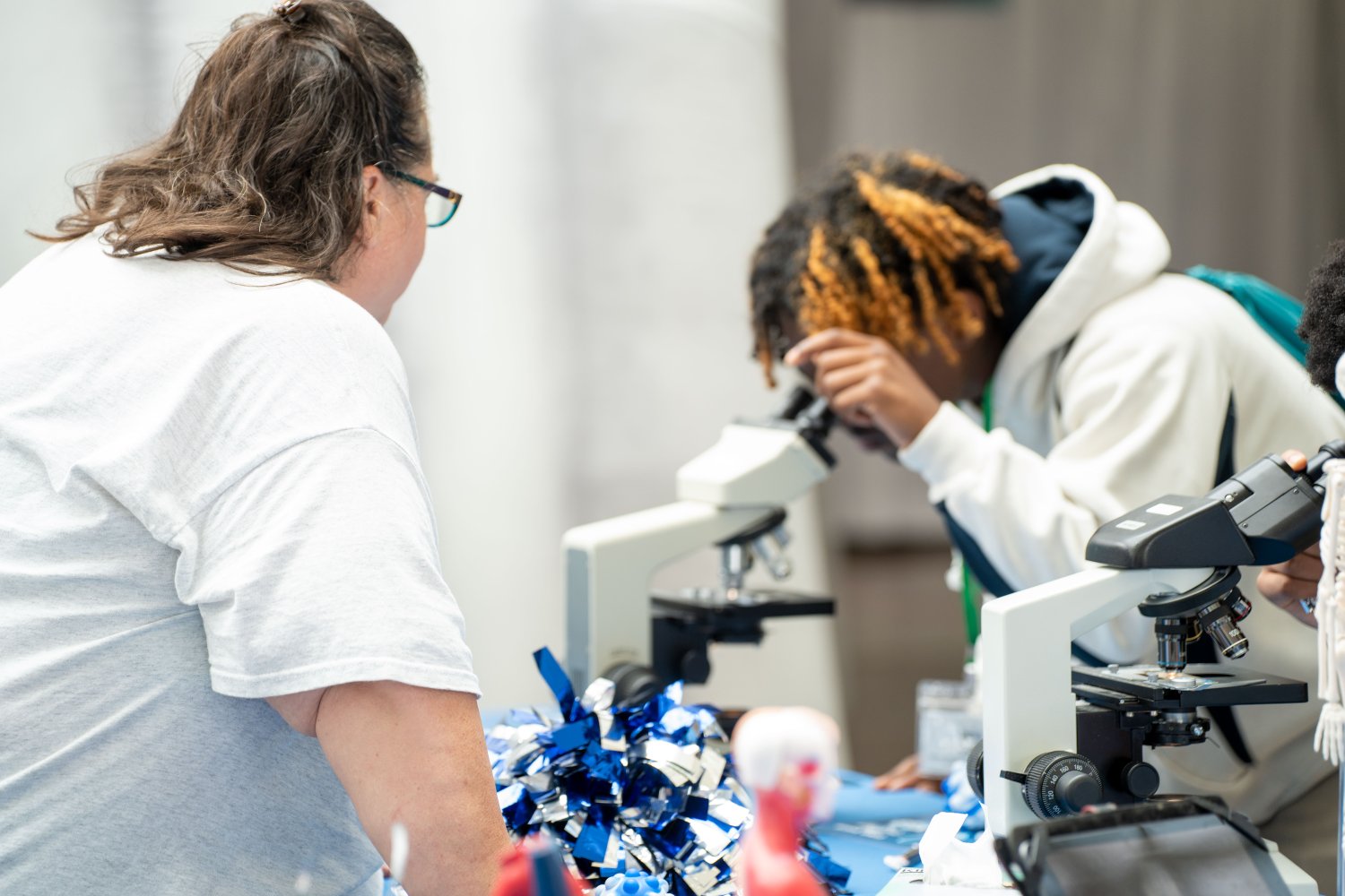 A close-up of students looking into microscopes at a healthcare education booth. The foreground shows anatomical models and blue-and-white pom-poms. One student wears a white hoodie with a green lanyard and another uses a separate microscope. A person in a white graphic t-shirt is visible in the left foreground, assisting or supervising.
