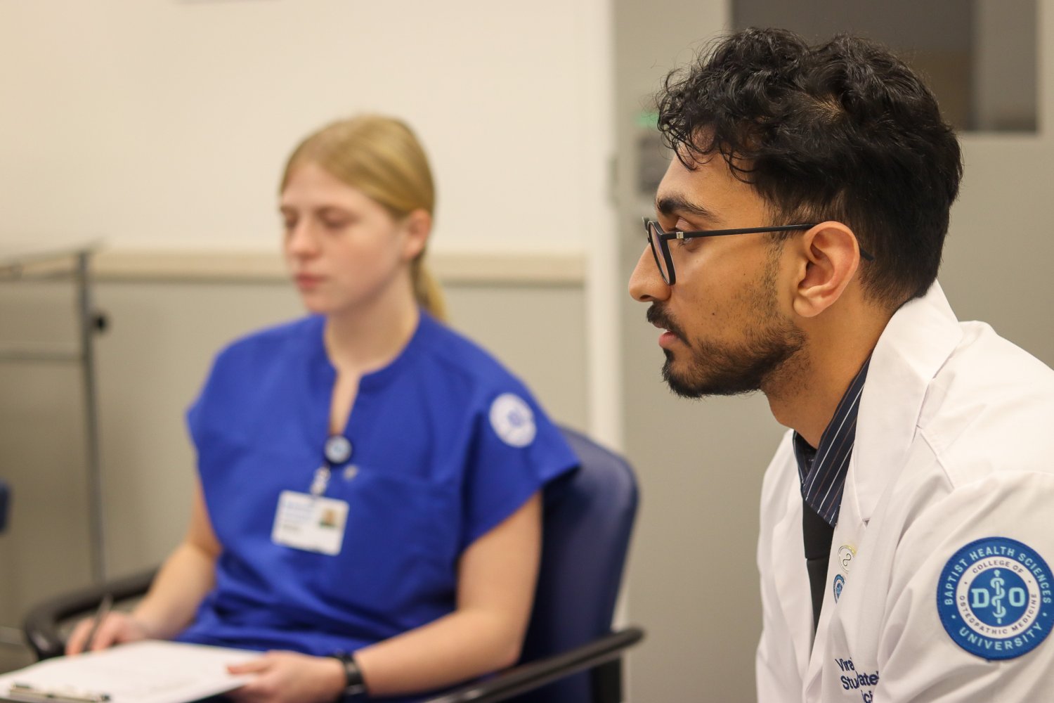 A nursing student in blue scrubs and a medical student in a white coat sit in a classroom during an interprofessional simulation. The medical student is listening attentively, while the nursing student holds a clipboard and takes notes.