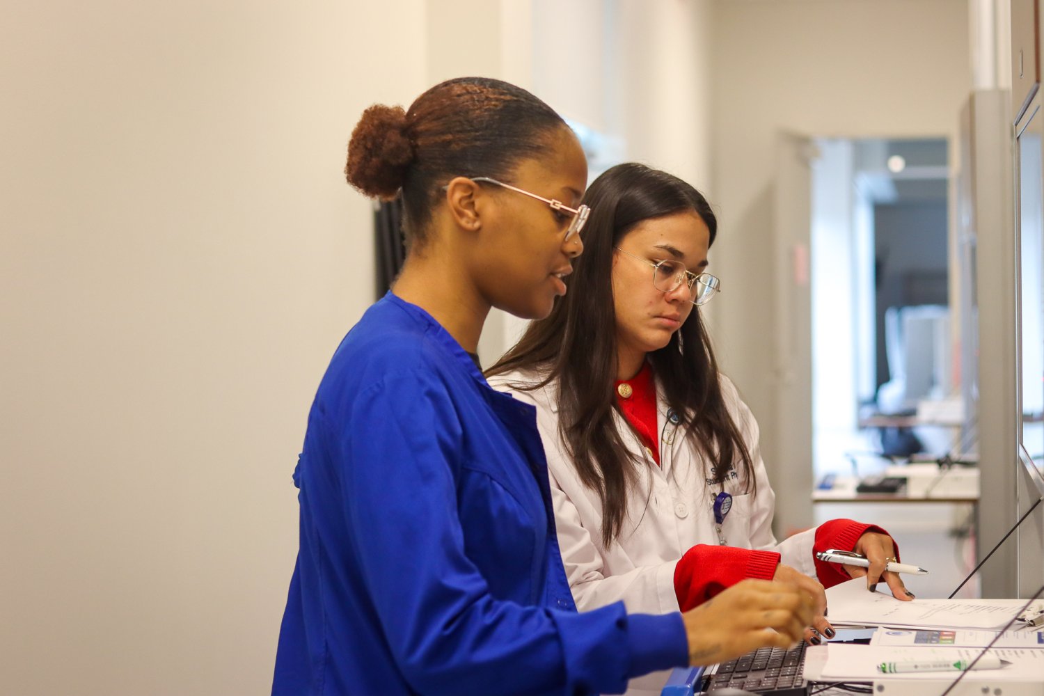 A nursing student in blue scrubs works alongside a medical student in a white coat at a computer station. Both students are focused on reviewing documents and entering information during the simulation lab activity.