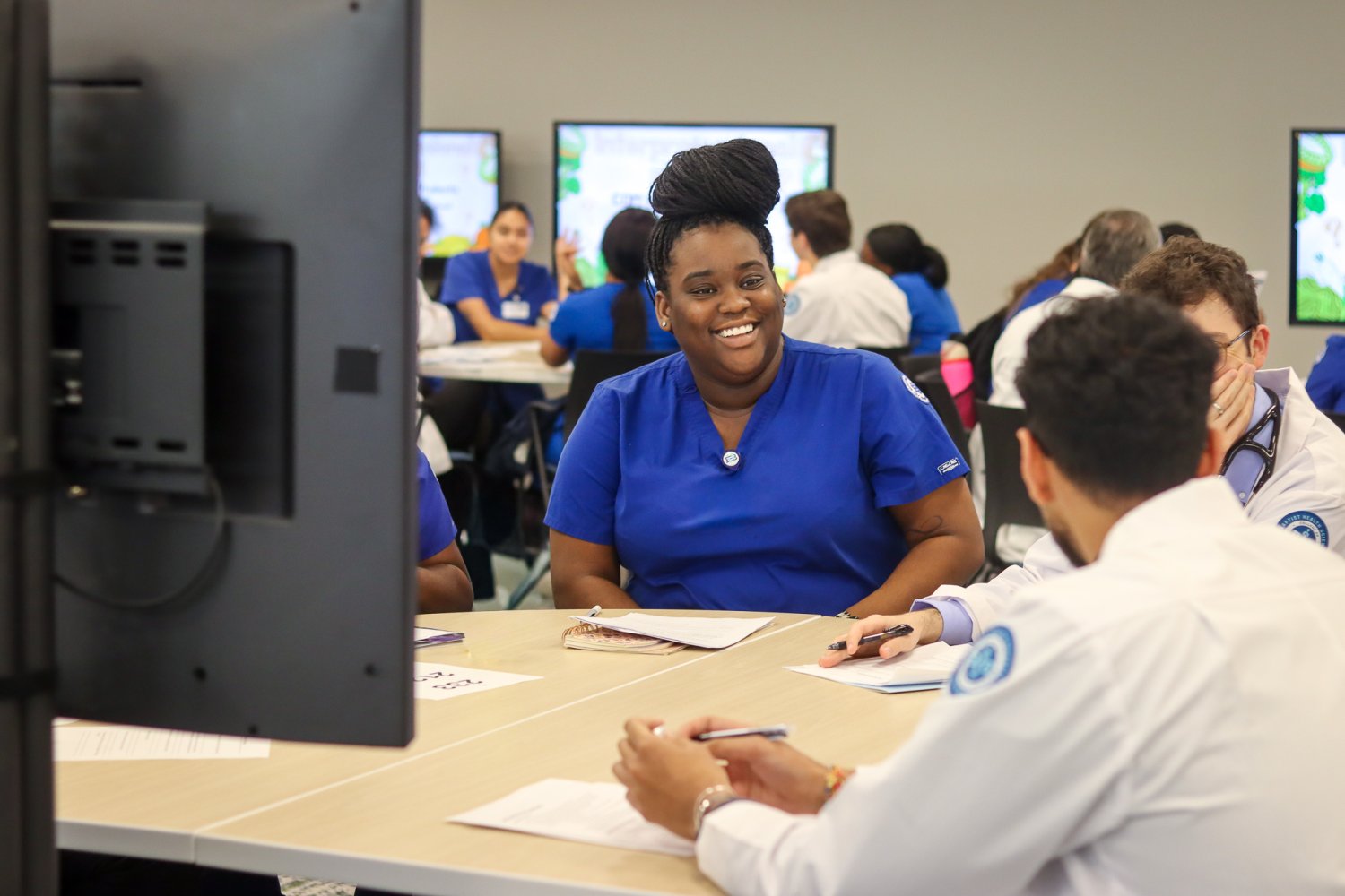 A nursing student in blue scrubs smiles while sitting at a table with medical students in white coats during a group debrief session. Other student groups are visible working together in the background.