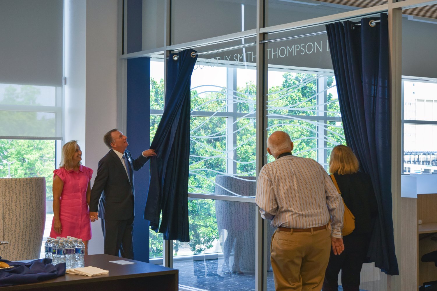 A man unveils a curtain while holding hands with a woman in a bright pink dress during the dedication of "The Dorothy Smith Thompson Library." Two other individuals watch as the curtain is pulled back, revealing the name etched on the glass.