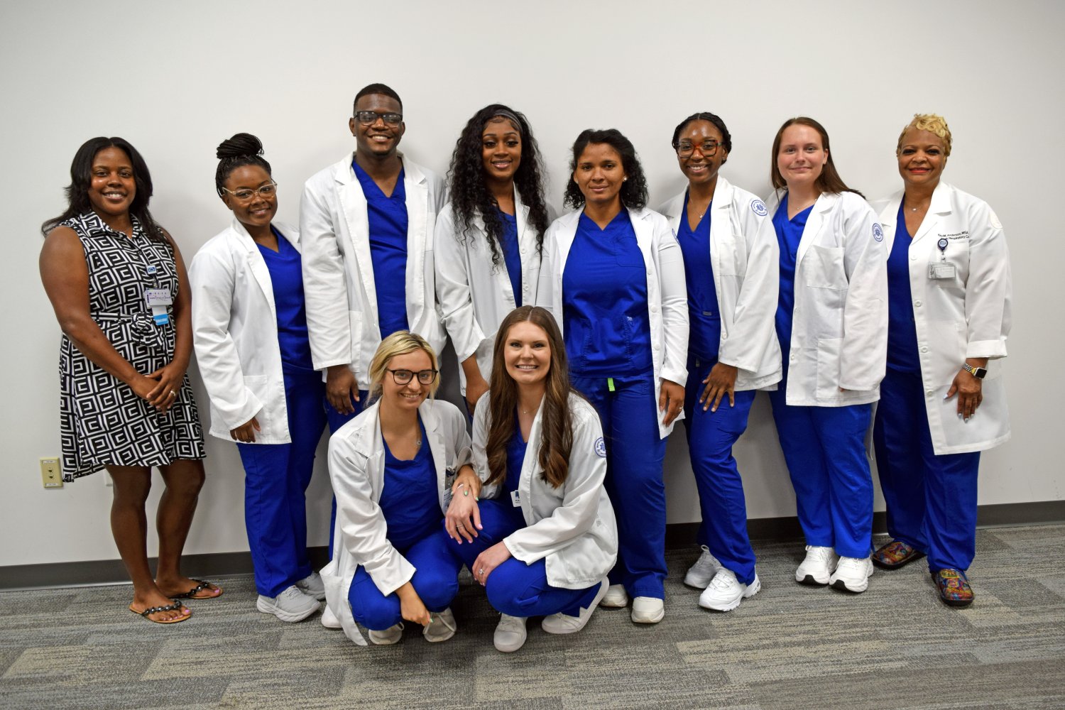 group of students in blue scrubs and white coats standing in front of a blank wall with professors