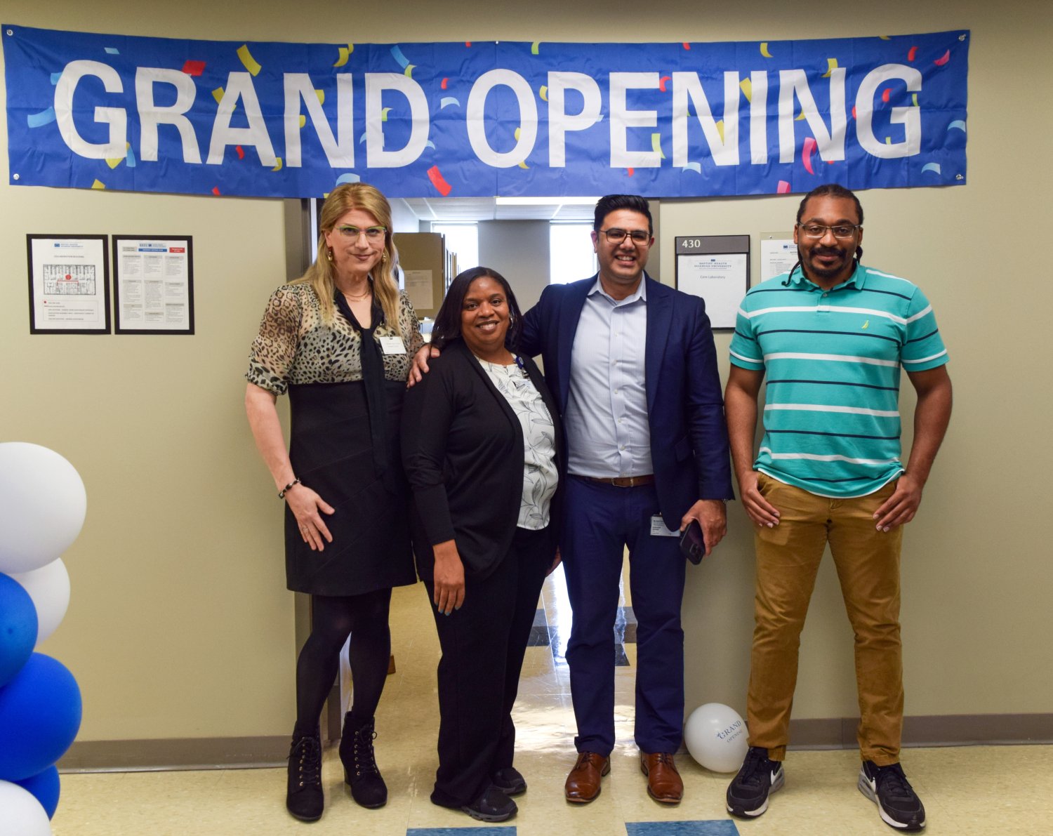 grand opening sign on the wall and blue and white balloons with four people in the photo and two people cutting a blue ribbon