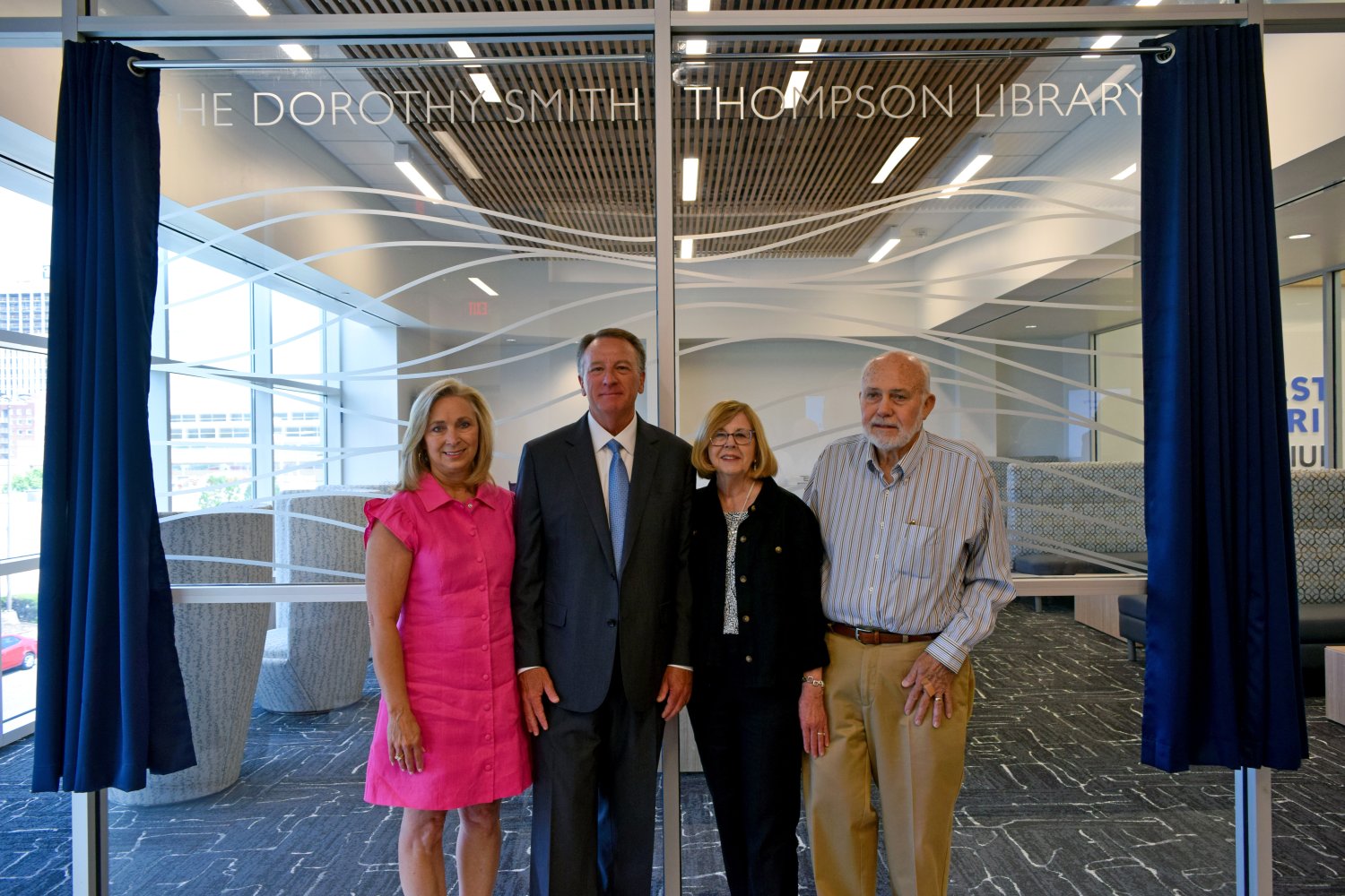 Four people pose in front of the glass wall of "The Dorothy Smith Thompson Library." The group includes a woman in a pink dress, a man in a suit and blue tie, a woman in black with glasses, and another man in a striped shirt and khakis.