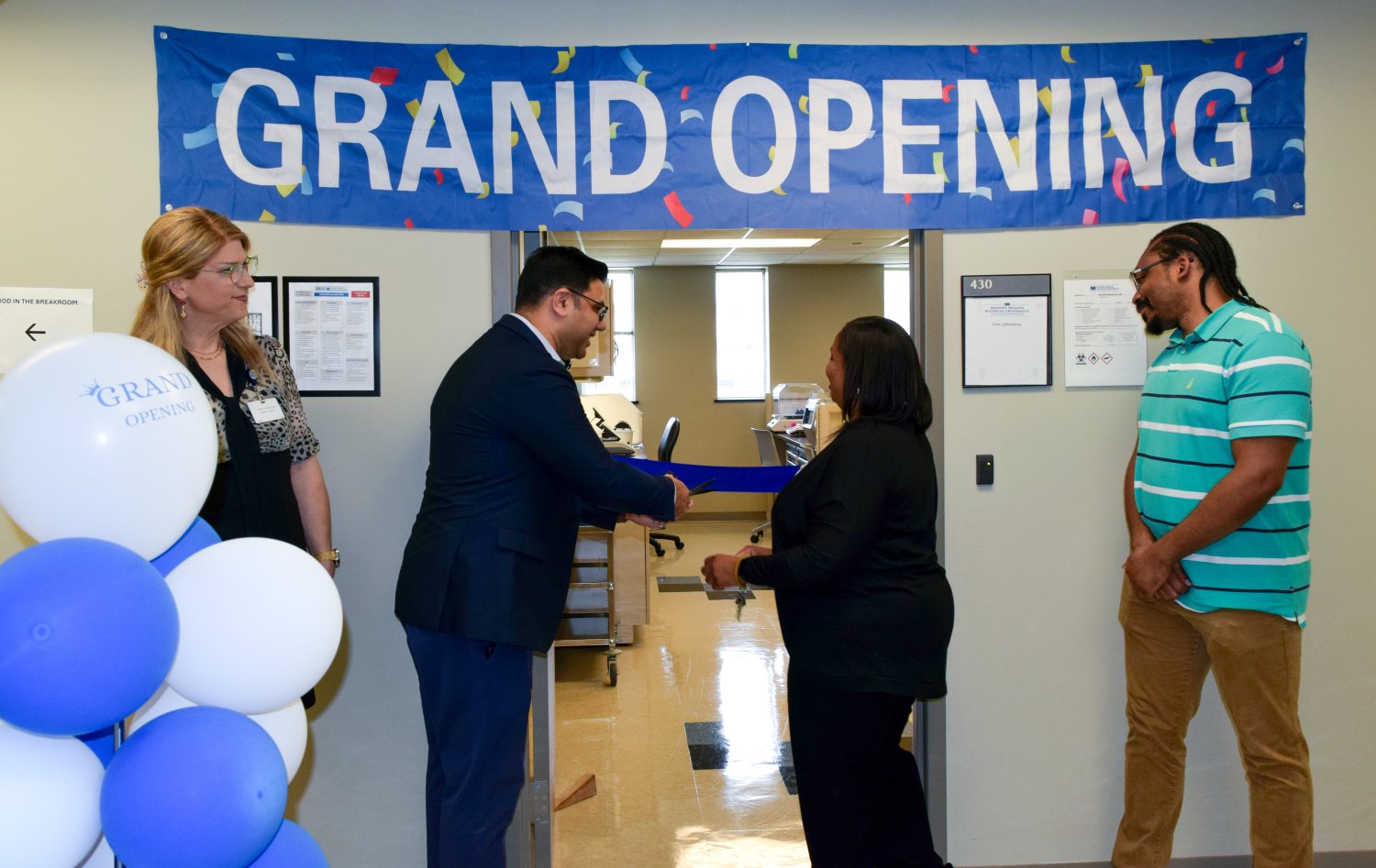 grand opening sign on the wall and blue and white balloons with four people in the photo and two people cutting a blue ribbon