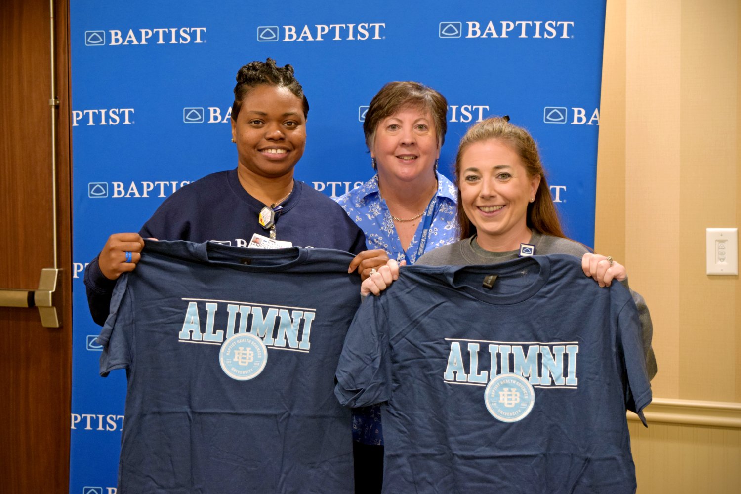 Three BHSU alumni smile and proudly display their alumni T-shirts in front of a Baptist-branded backdrop.