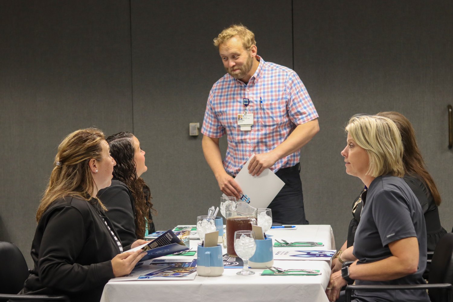 A man in a plaid shirt stands at a table engaging with four seated attendees during a lunch-and-learn event. The table is set with informational materials, mugs, and iced drinks.