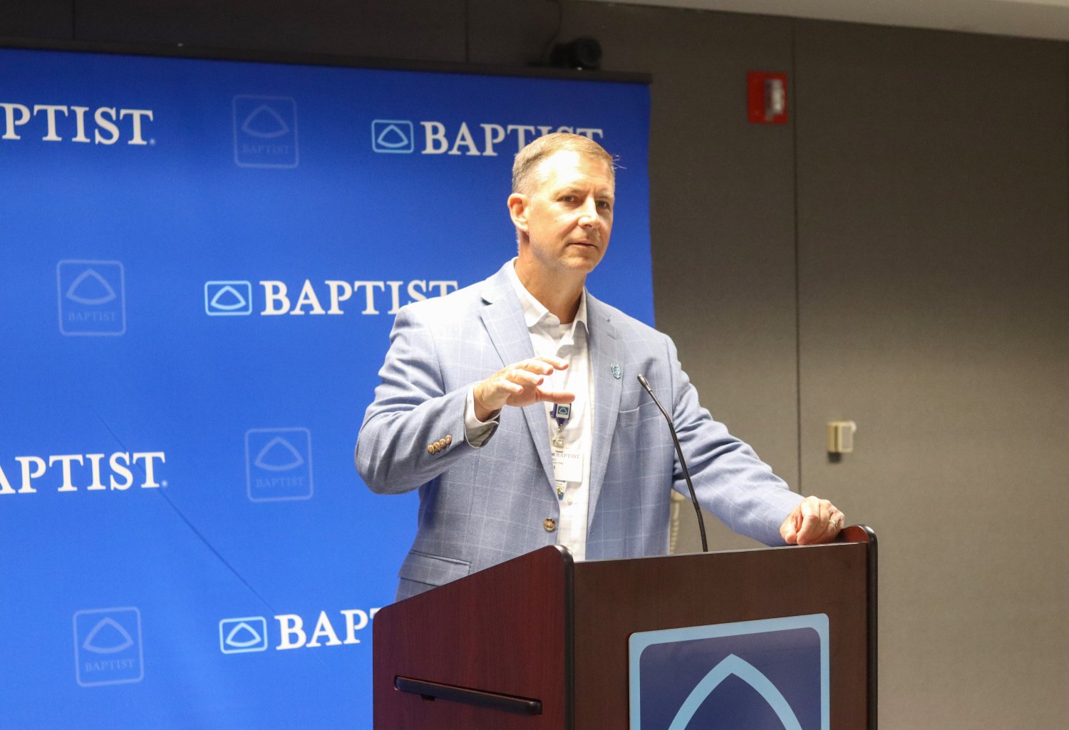 A speaker stands at a podium branded with the Baptist logo, addressing the audience at a lunch-and-learn event. A blue Baptist backdrop is visible behind him.