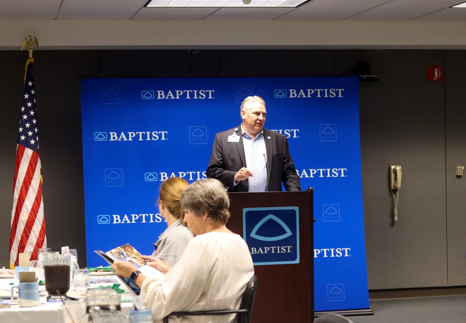 A man in a blazer speaks at a podium in front of an American flag and Baptist-branded backdrop, addressing attendees seated at tables.