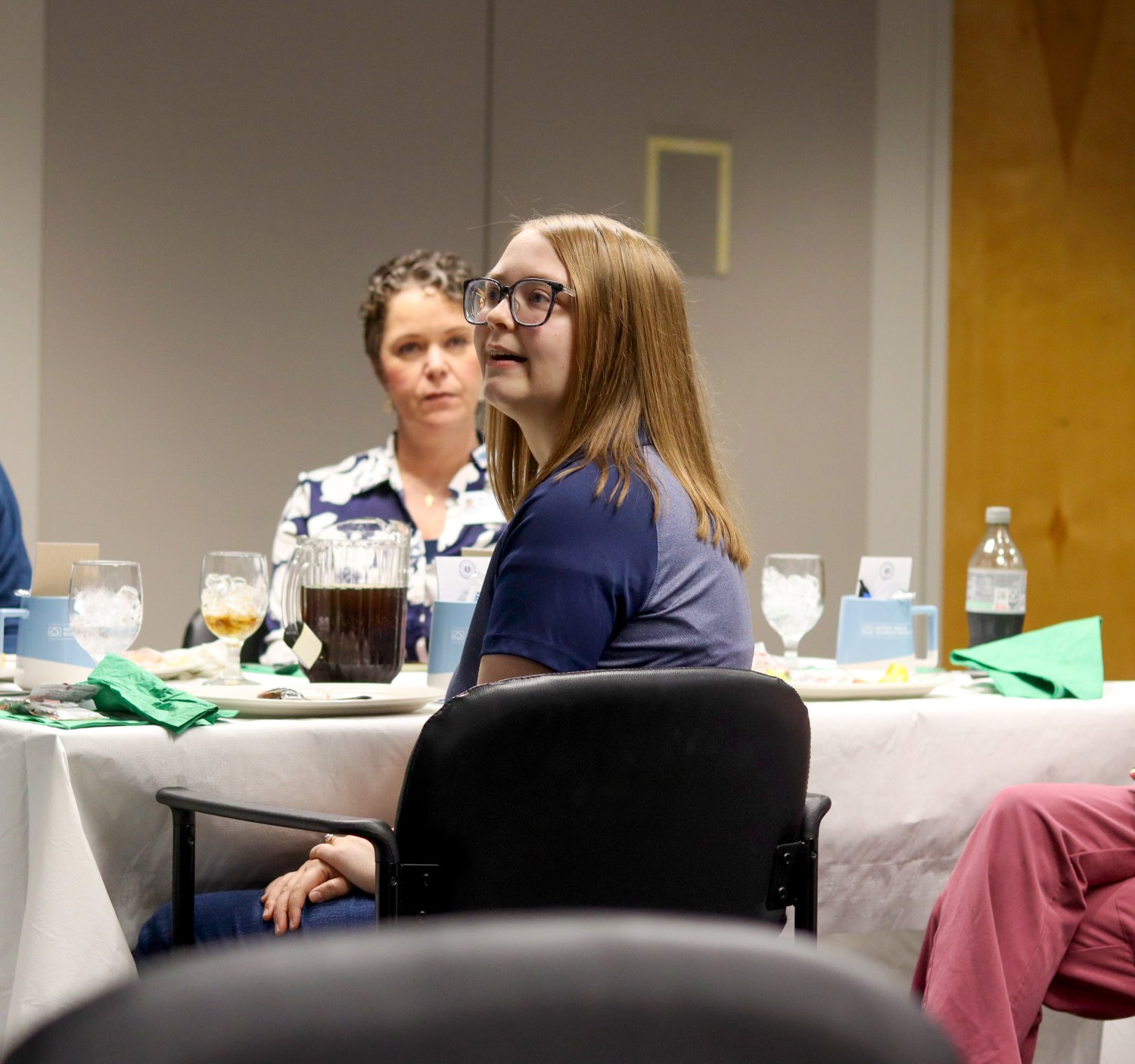 A young woman with glasses listens attentively at a lunch table set with drinks and materials. Another attendee is seated beside her.