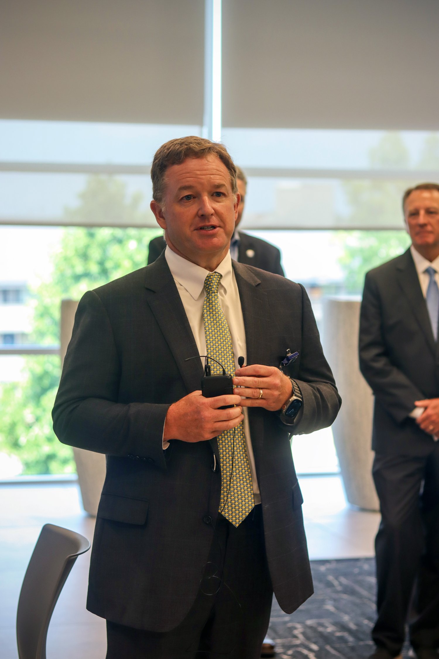 A man in a suit speaks while holding a microphone, with others standing behind him in a modern, well-lit room. He appears to be giving remarks during a formal event or ceremony.