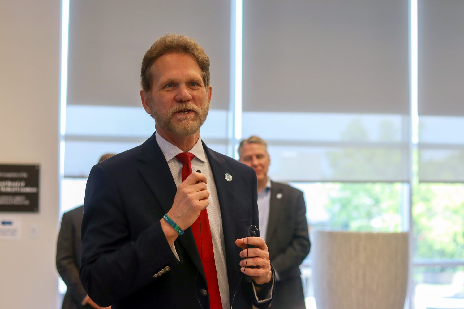 A man with a beard and red tie speaks into a microphone, holding his glasses in one hand. He stands in front of a seated audience in a professional setting with others behind him listening attentively.