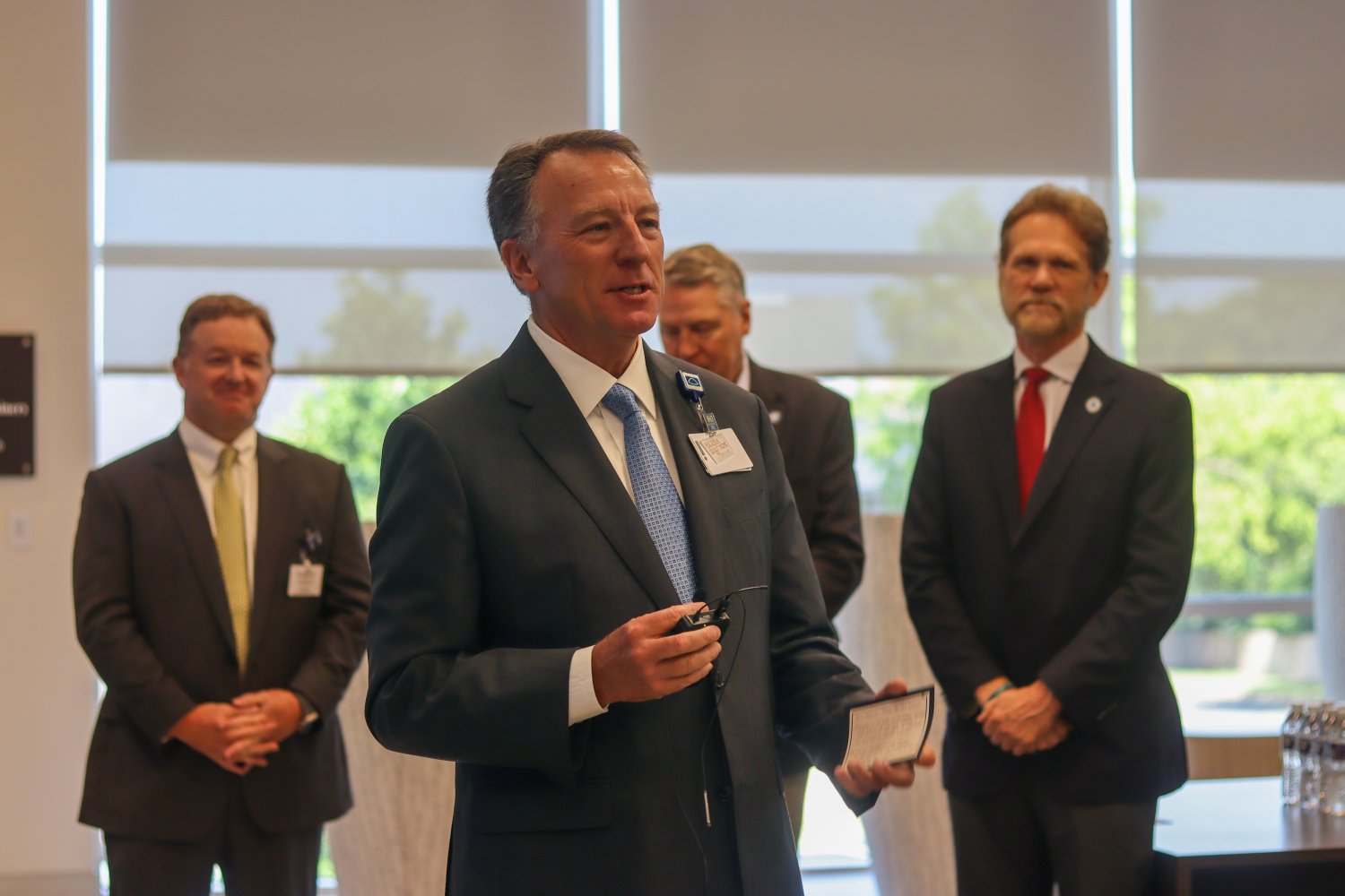 A man in a dark suit and blue tie speaks to an audience, holding a notecard and a lapel microphone. Several individuals in suits stand behind him in support.