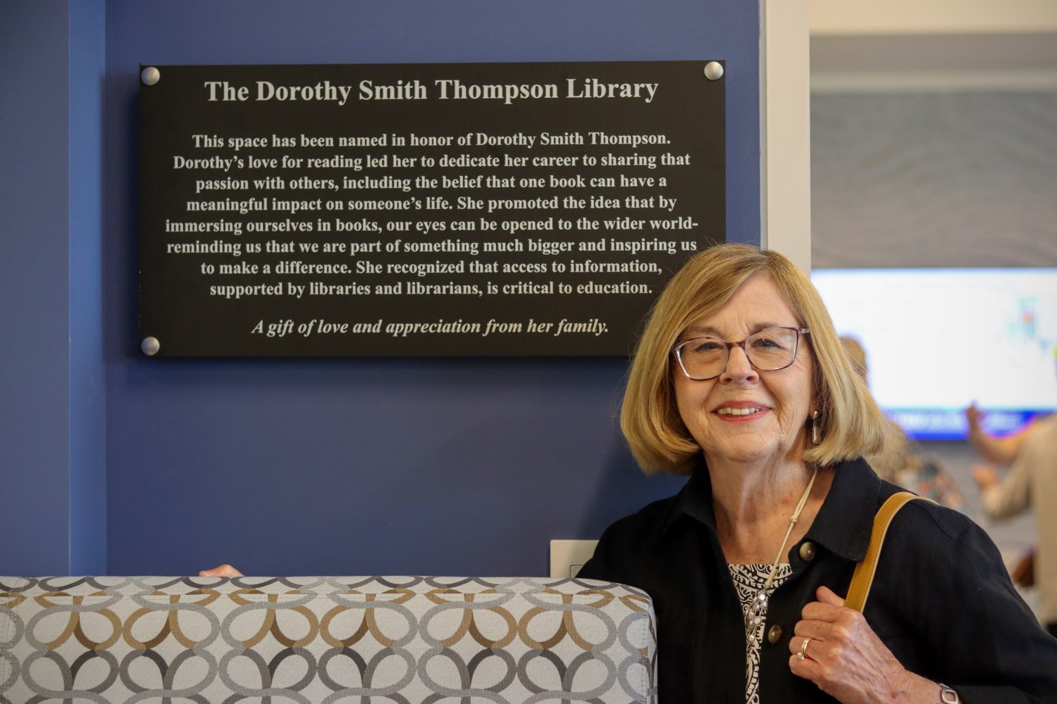 A woman smiles while standing next to a wall-mounted plaque titled "The Dorothy Smith Thompson Library." The plaque explains the dedication in her honor, highlighting her love for reading and contributions to education.