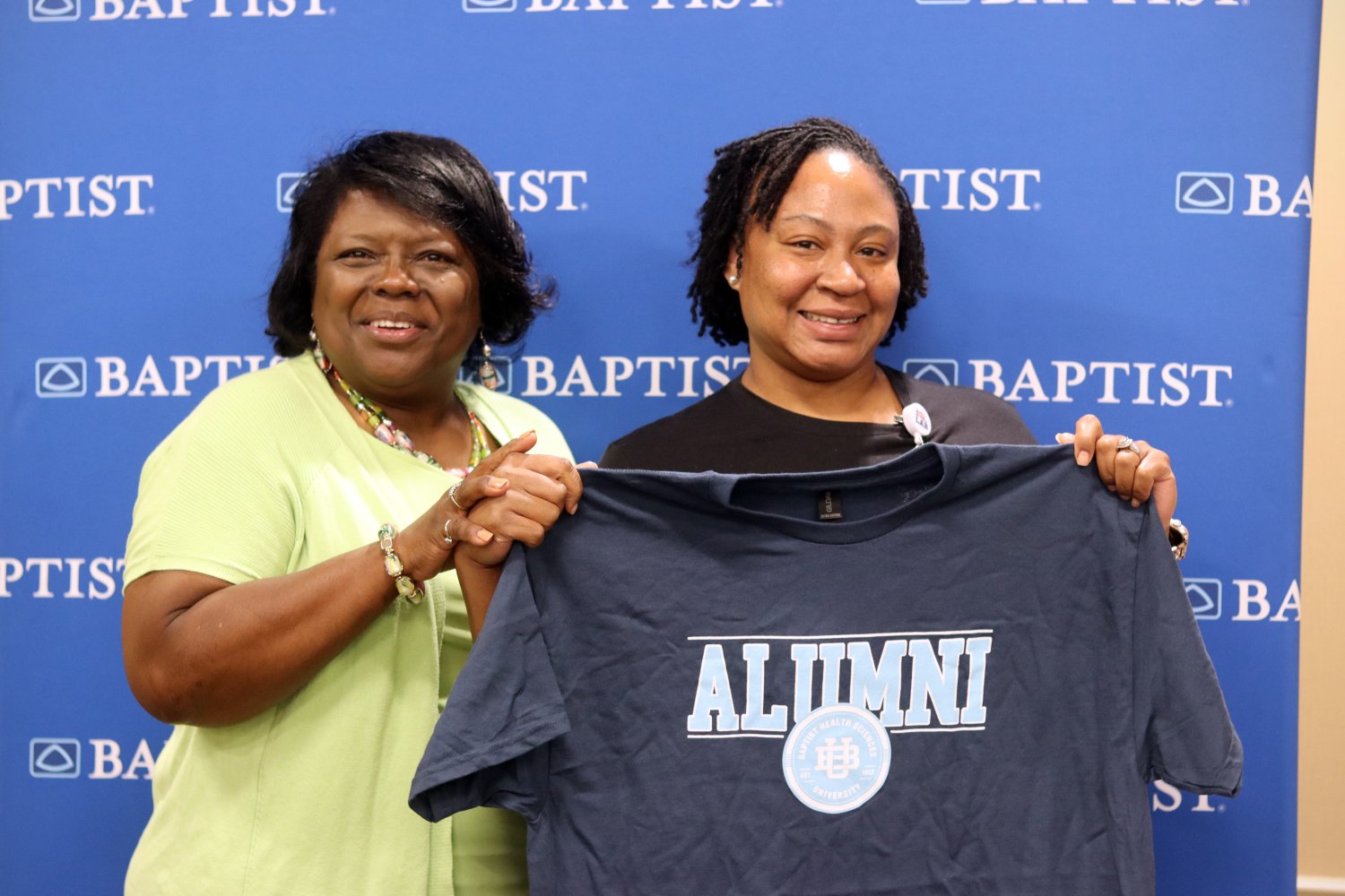 Two women smile and hold up a BHSU alumni T-shirt together, standing in front of a Baptist-branded step-and-repeat banner.