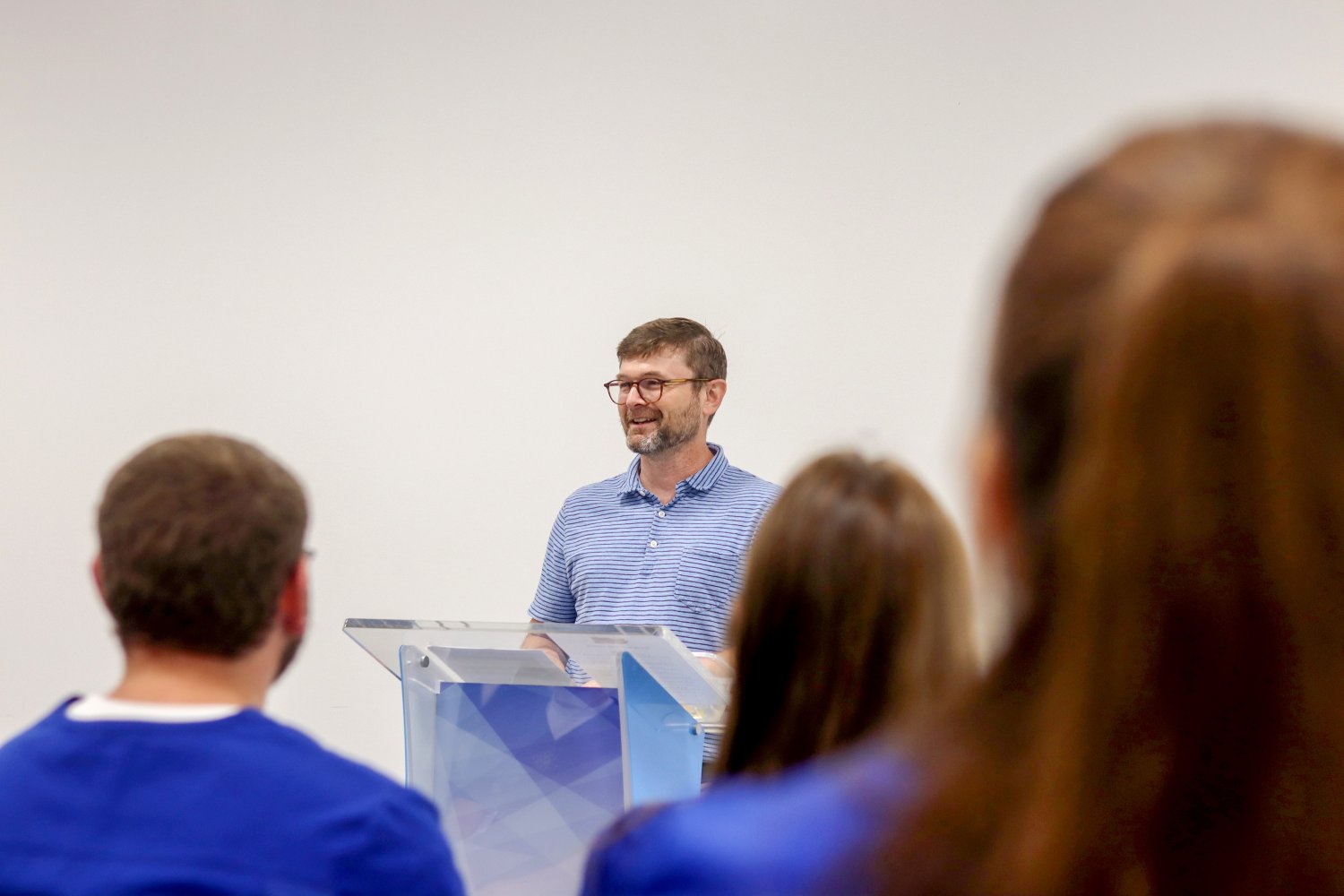 A man in a striped polo shirt stands at a clear podium, smiling as he speaks to an audience of students in blue scrubs.