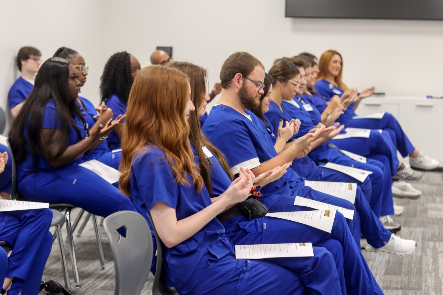 A group of students in blue medical scrubs sits in rows, clapping and smiling during a formal ceremony.