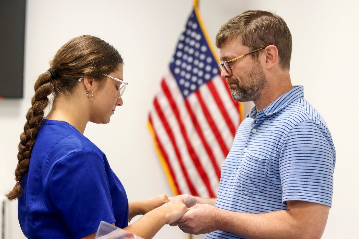 A male speaker and a female student in blue scrubs share a solemn moment, holding hands in front of an American flag.