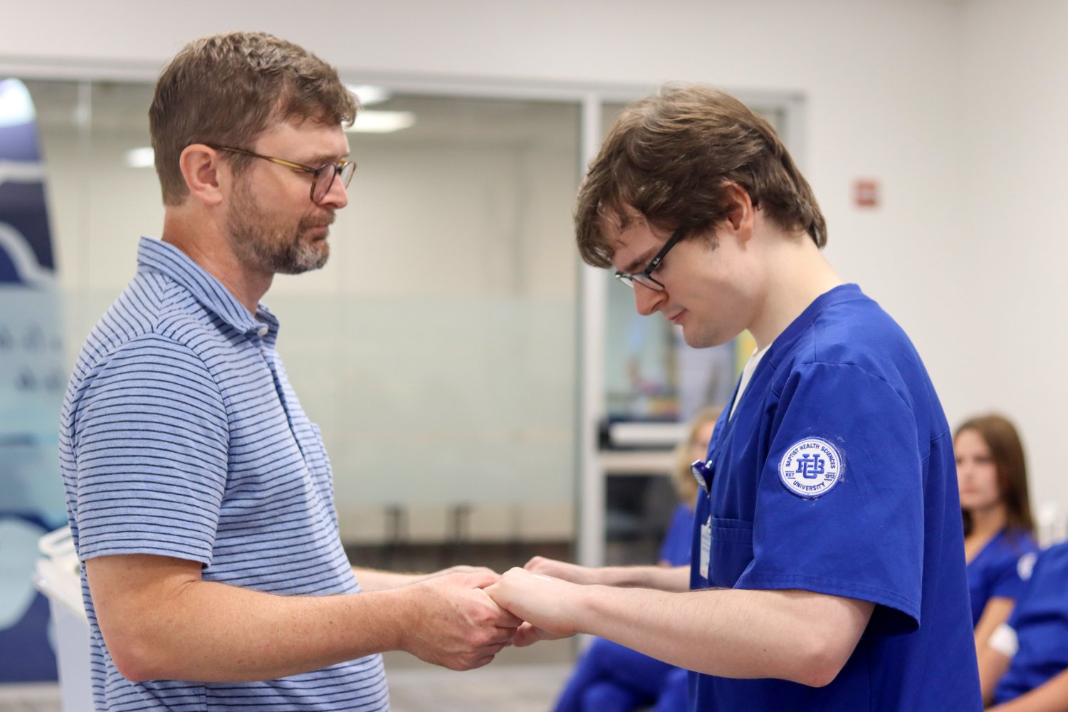 A man and a student in blue scrubs share a focused and quiet moment during a ceremonial hand blessing.